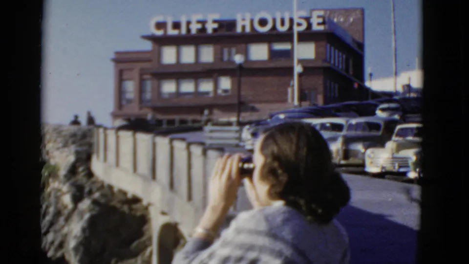 The woman continues looking through binoculars at the Cliff House overlook, framed by the visible sign, railing, rocky edge, and classic cars.