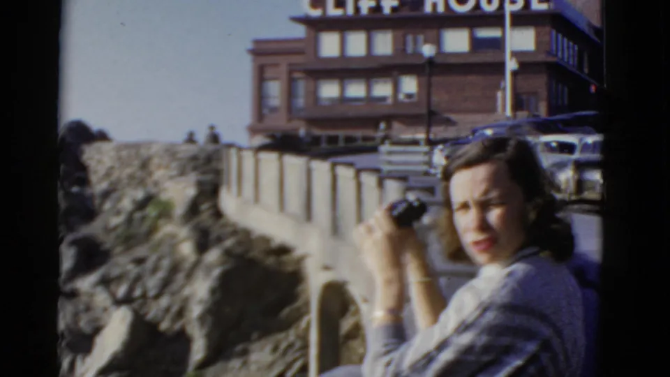 At the Cliff House overlook in San Francisco, the woman turns toward the camera while holding binoculars, with parked cars and the building behind her.
