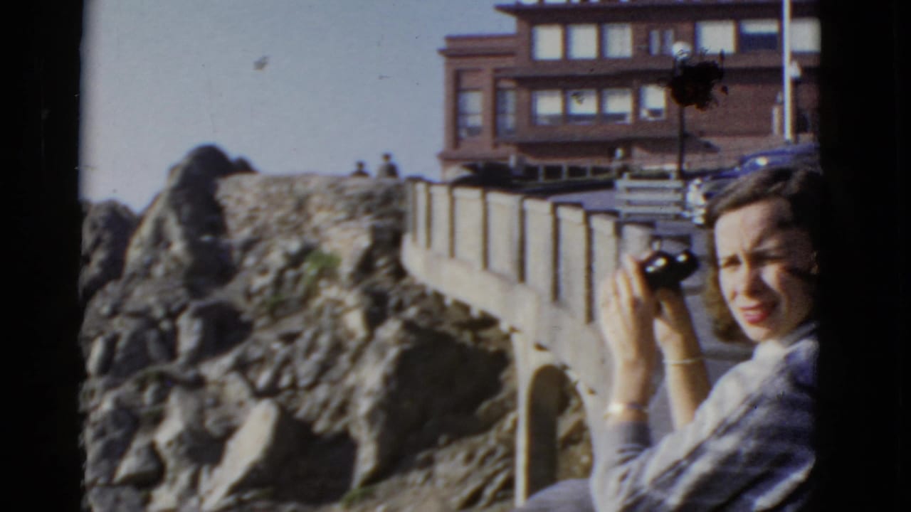 In 1952 San Francisco home-movie footage, a woman with binoculars stands at a stone coastal overlook near rocky cliffs and a cliffside building.