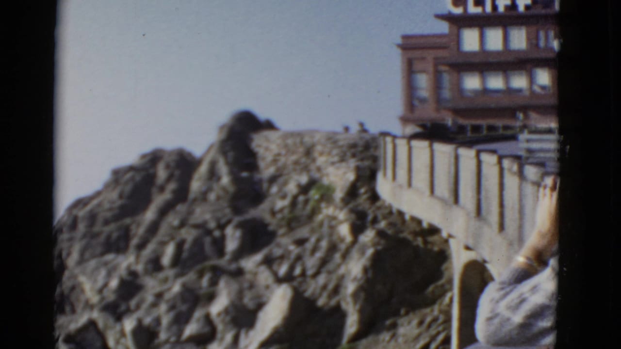 The San Francisco coastal overlook remains in view, with rugged rock, a stone railing, partial building signage, and the woman cropped at the edge.
