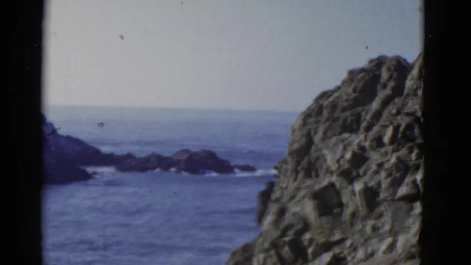 Vintage 8mm San Francisco coastal view with a large rock face at right and dark offshore formations across the water.