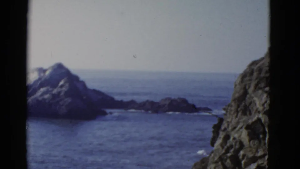 1952 home-movie frame near San Francisco showing a jagged island, low rocky outcrops, open water, and a cliff edge at right.