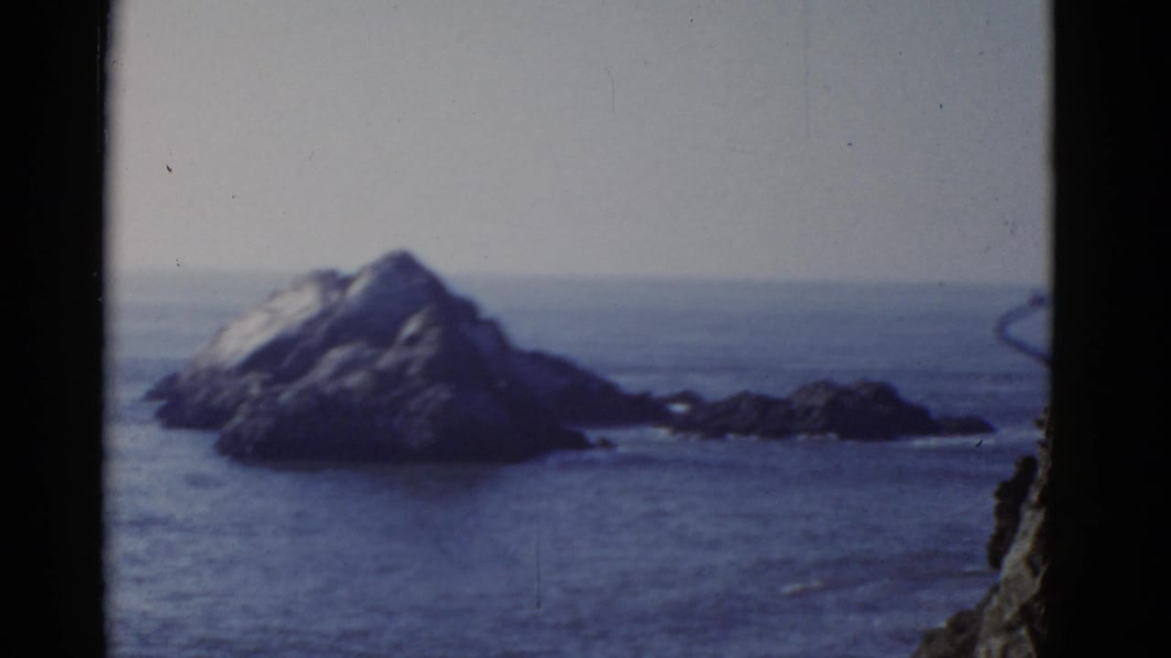 1952 San Francisco-area 8mm frame showing a rocky offshore formation, blue water, pale sky, and dark film-gate edges.