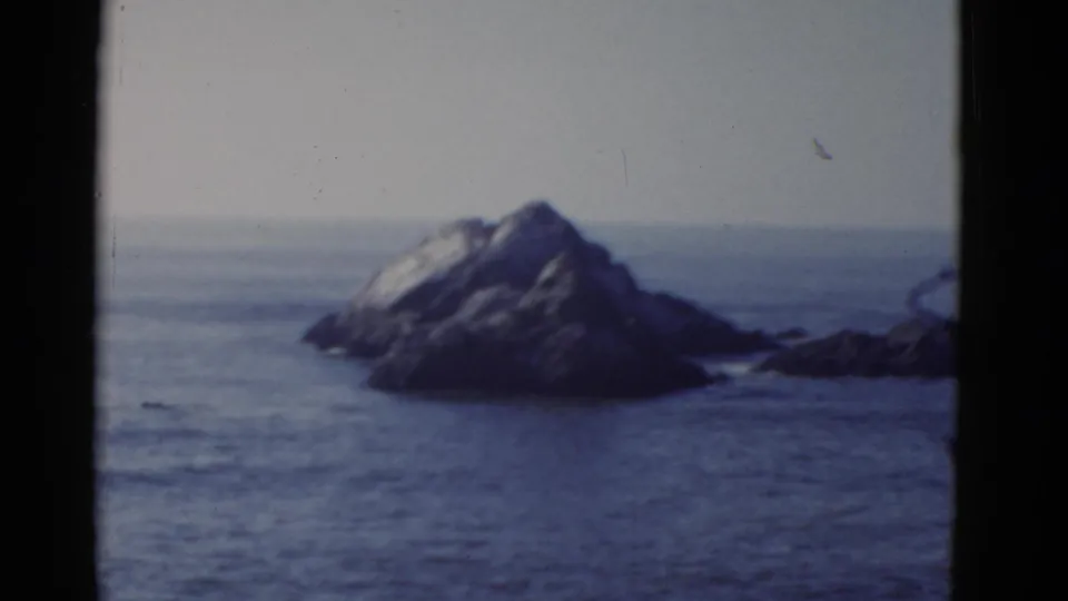 1952 San Francisco-area 8mm footage shows a soft-focus rocky formation in blue ocean under a washed pale sky.
