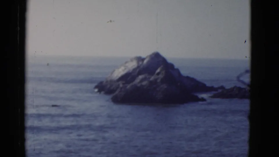 1952 San Francisco-area home-movie frame with a rocky sea stack, blue ocean, faint horizon, and aged 8mm film texture.
