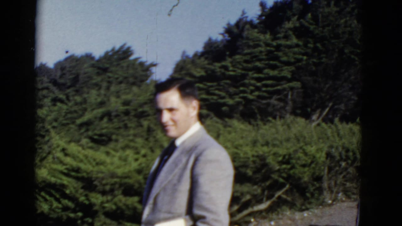Adult man in a light gray suit standing near dense green shrubs and trees.
