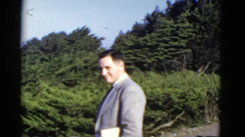 The 1952 8mm frame shows a suited adult man in profile near thick shrubs and trees, with soft home-movie focus.