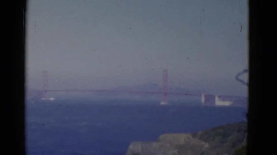 1952 Bay Area 8mm frame showing a hazy, distant Golden Gate Bridge with blue water and foreground coastal terrain.