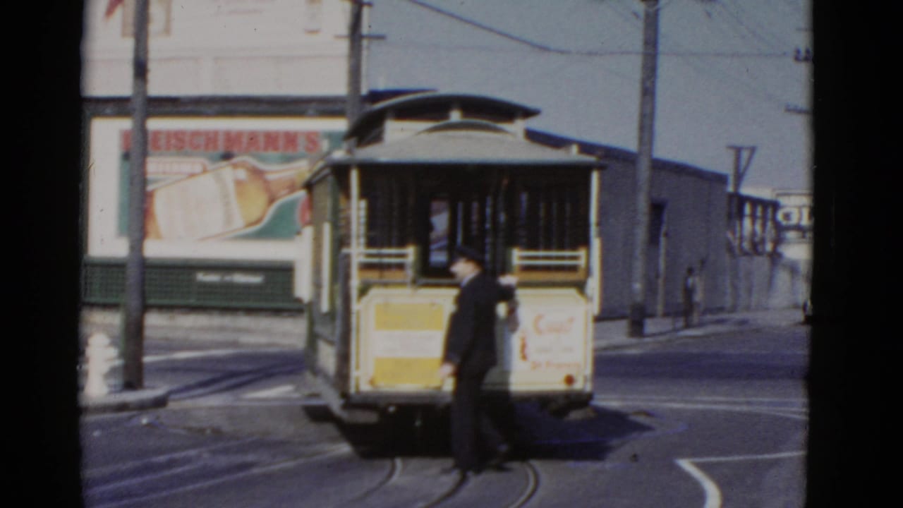 Conductor stands near the back of a cable car on curved street rails.
