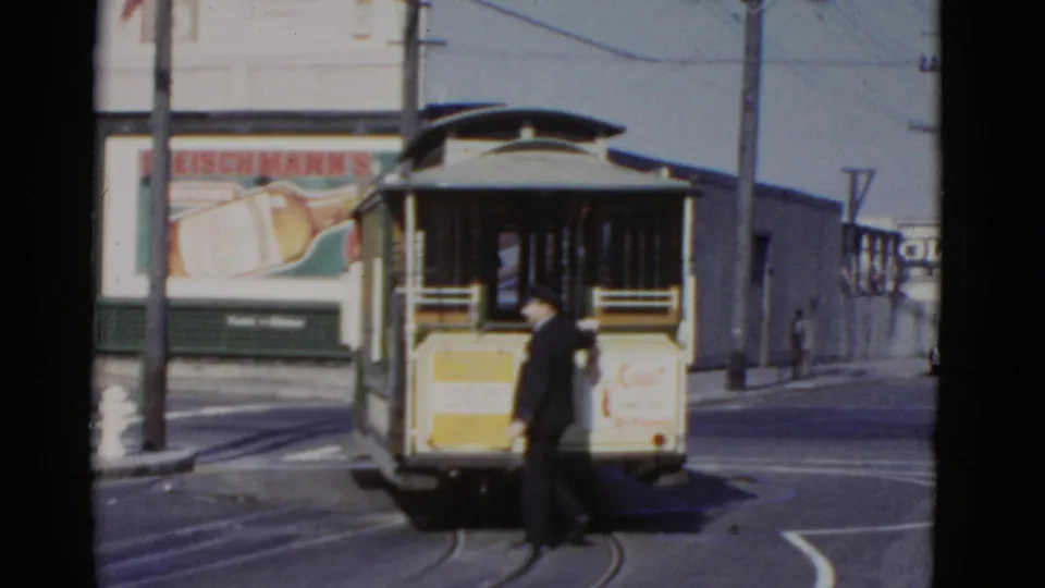 A uniformed conductor stands beside the rear of a San Francisco cable car, with rails, overhead wires, and billboard signage behind him.