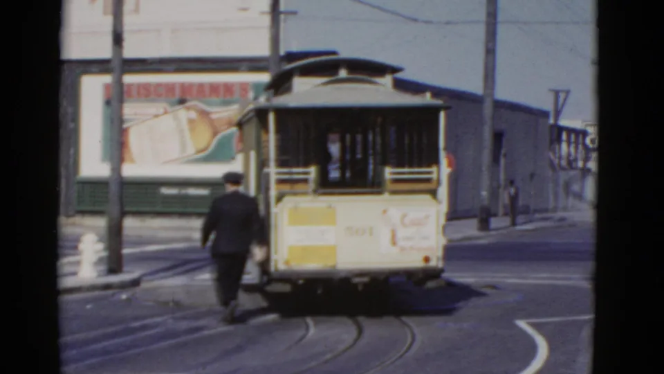The conductor walks away from the camera behind a vintage cable car on San Francisco street rails, with billboard advertising in the background.