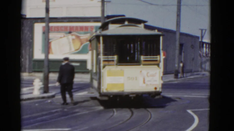 A conductor in dark uniform follows the rear of a cable car along curved rails in a San Francisco street scene from supplied 1952 footage.