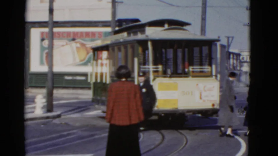 Woman in a red plaid jacket stands near a 1952 San Francisco streetcar as pedestrians and a uniformed worker appear around the tracks.
