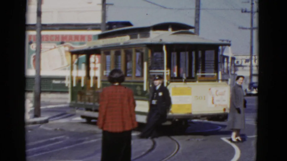 1952 San Francisco streetcar scene with a uniformed transit worker beside the car, pedestrians nearby, rails, wires, and billboard signage.