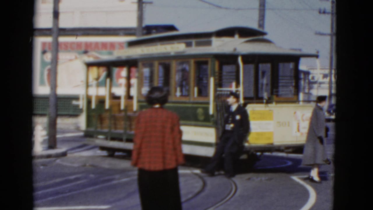 1952 San Francisco street view with a green and cream cable car turning while a woman in a red plaid coat watches.