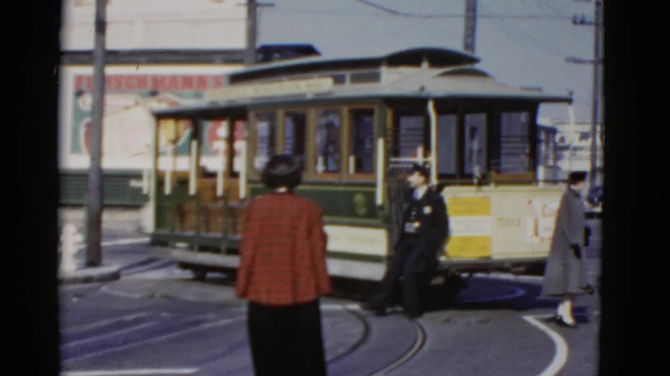 1952 San Francisco street view with a green and cream cable car turning while a woman in a red plaid coat watches.