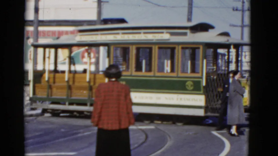 A side view of the 1952 San Francisco cable car fills the frame as a woman in a red plaid coat watches from the street.