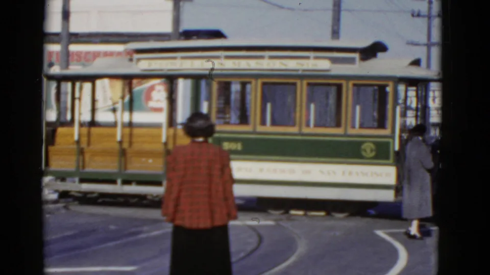 The green and cream cable car completes more of its turn on a San Francisco street, with pedestrians and storefront signage behind it.
