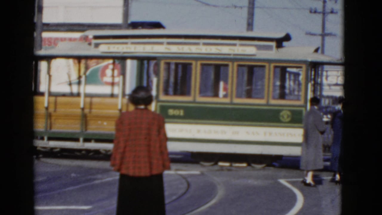 1952 San Francisco home-movie frame showing a green and white rail car behind waiting pedestrians and curved street tracks.