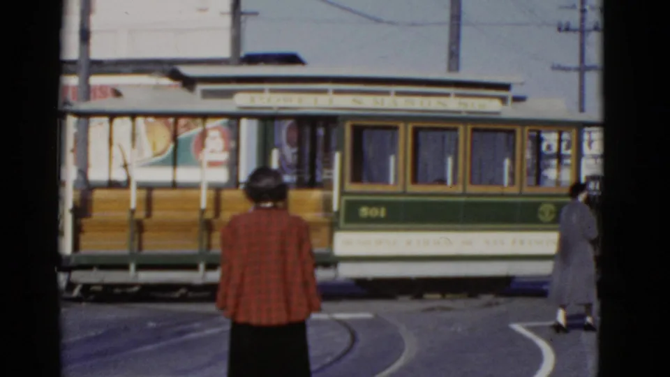1952 San Francisco street scene showing the side of a green and white rail car on tracks with pedestrians nearby.
