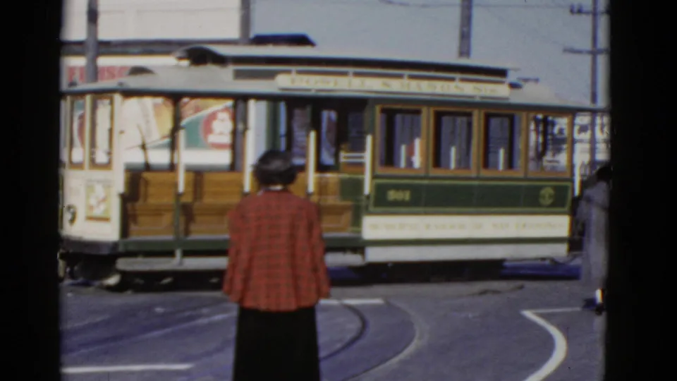 1952 San Francisco home-movie frame of a green and white rail car turning on tracks as pedestrians stand near the curb.