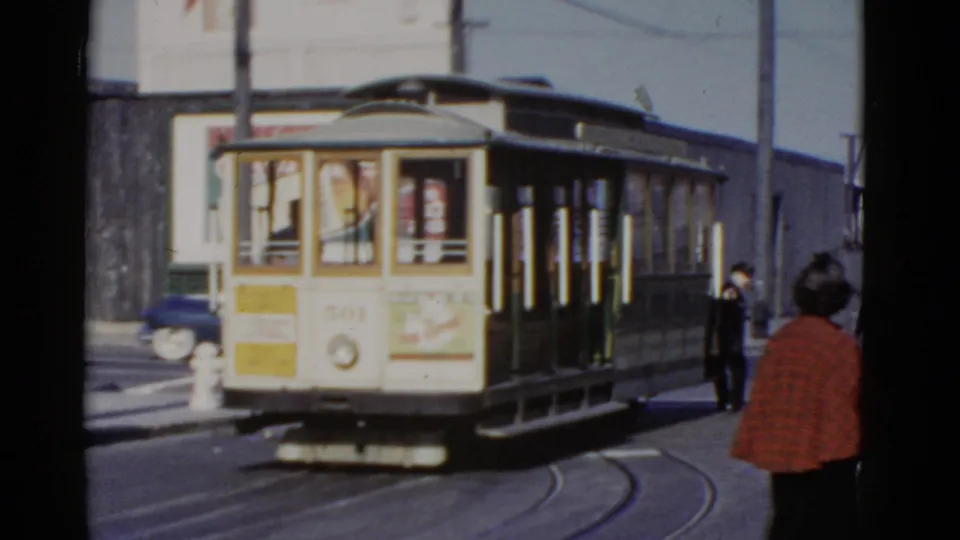 San Francisco cable car travels away along curved rails, with a woman in a red plaid jacket at the edge of the frame.