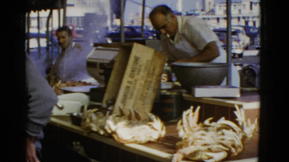 A seafood vendor works over a sidewalk stall in 1952 San Francisco footage, with crabs displayed and street traffic behind.