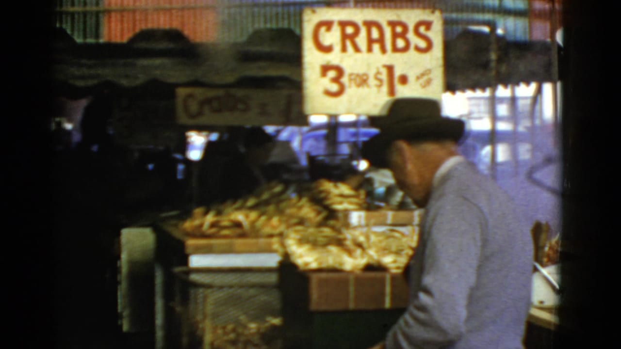In 1952 San Francisco market footage, a man in a fedora walks toward crates of crabs below a sign reading "CRABS 3 FOR $1."