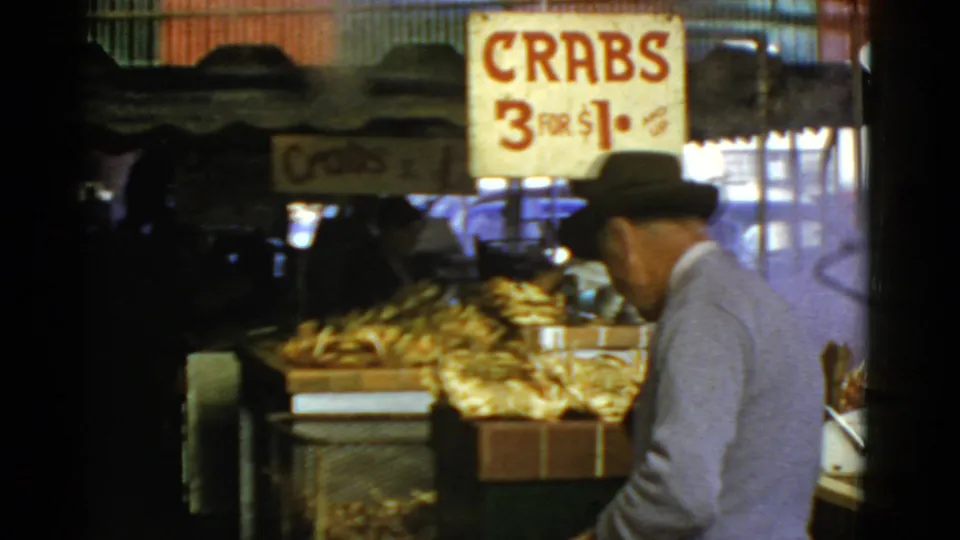 In 1952 San Francisco market footage, a man in a fedora walks toward crates of crabs below a sign reading "CRABS 3 FOR $1."