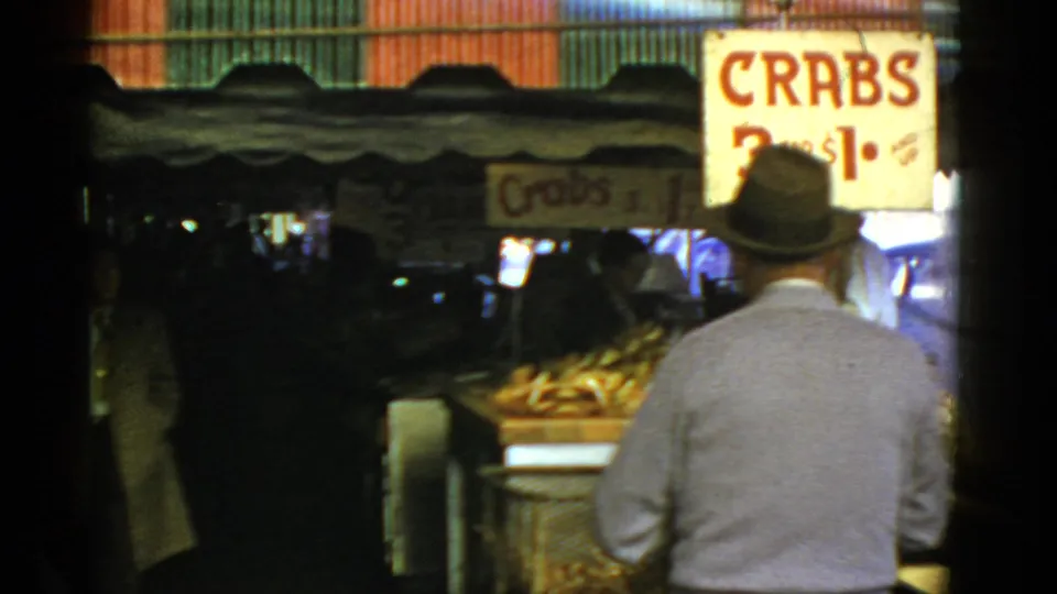 The passerby moves closer to a shaded crab vendor stall, with crates of seafood below the hand-painted price sign.