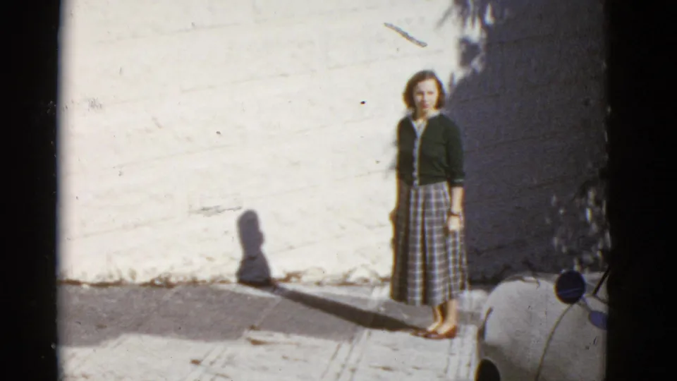 1952 San Francisco home-movie frame of a woman standing by a white wall, with her shadow on the sidewalk.