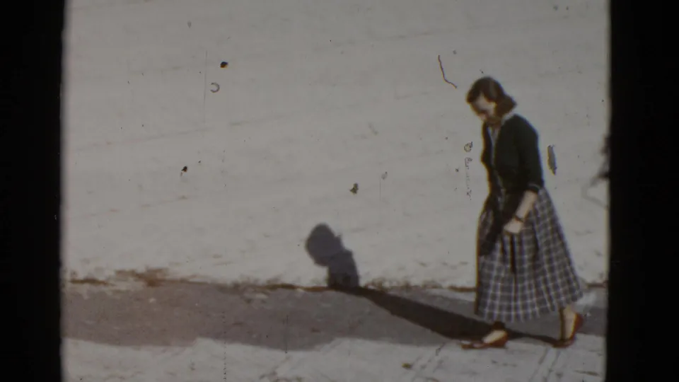 1952 San Francisco frame showing a woman in a dark top and plaid skirt walking beside a sunlit white wall.