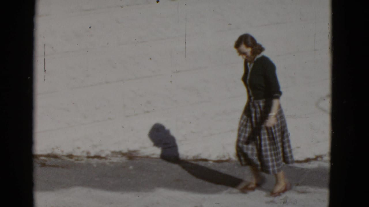 In supported 1952 San Francisco footage, a woman in a dark cardigan and plaid skirt walks along a bright sidewalk beside a plain wall.