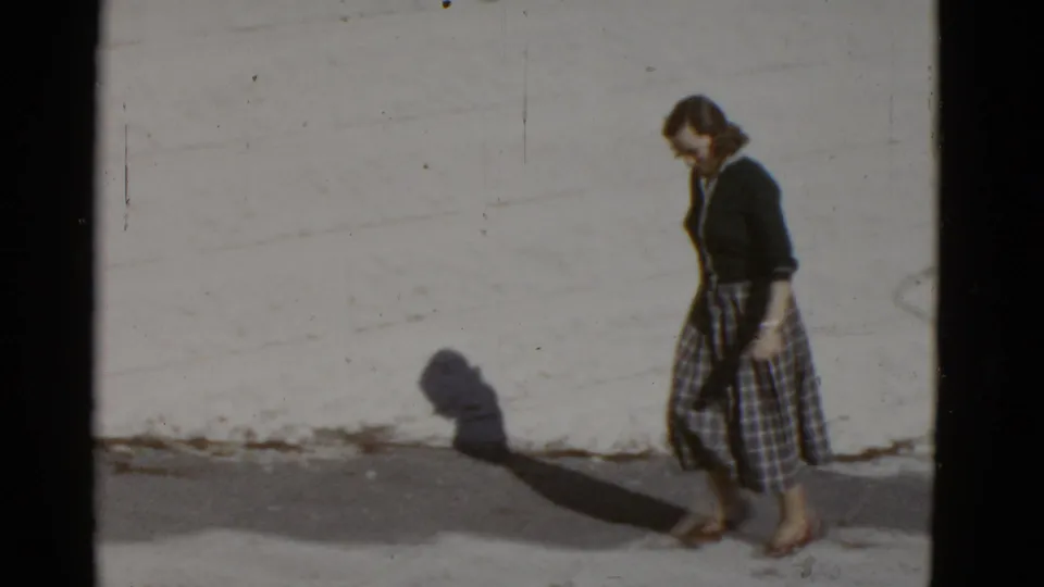 In supported 1952 San Francisco footage, a woman in a dark cardigan and plaid skirt walks along a bright sidewalk beside a plain wall.