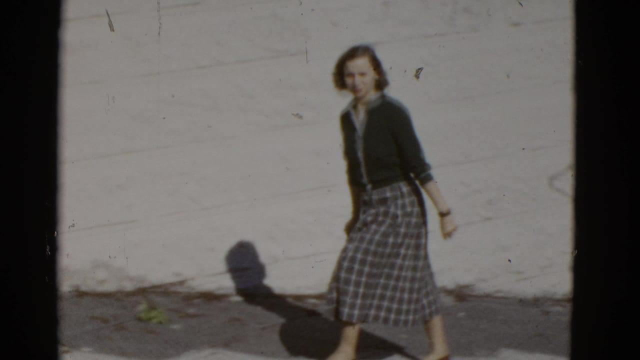 Woman glancing toward the camera while walking along a sidewalk beside a light wall.