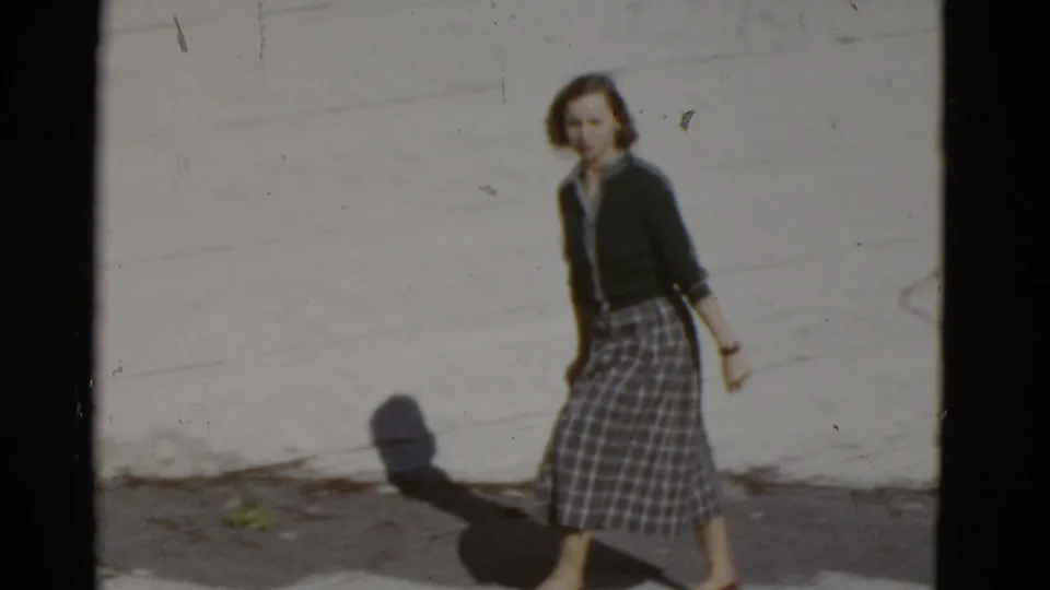 The woman turns toward the camera while walking past a pale wall, with strong sunlight casting her shadow on the pavement.