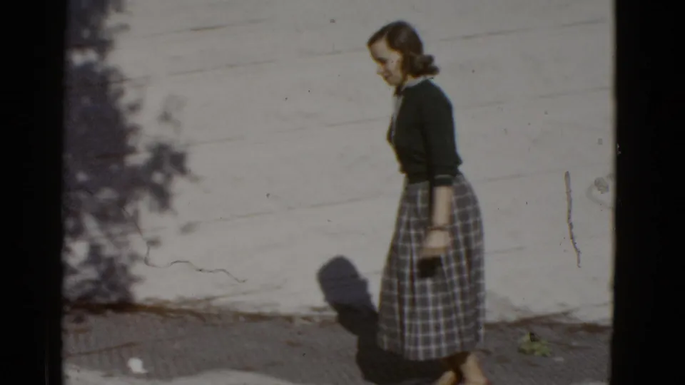 A side profile shows the woman continuing along the sidewalk, her plaid skirt and dark top framed against the pale wall.