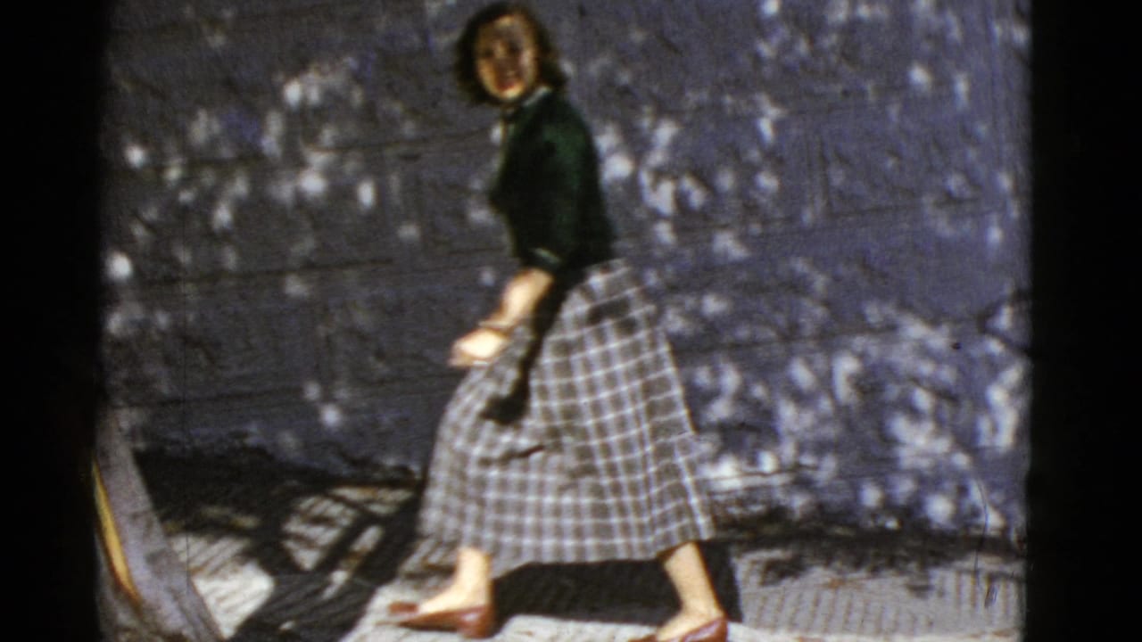 A woman in mid-century clothing walks beside a wall on a sun-dappled San Francisco sidewalk.