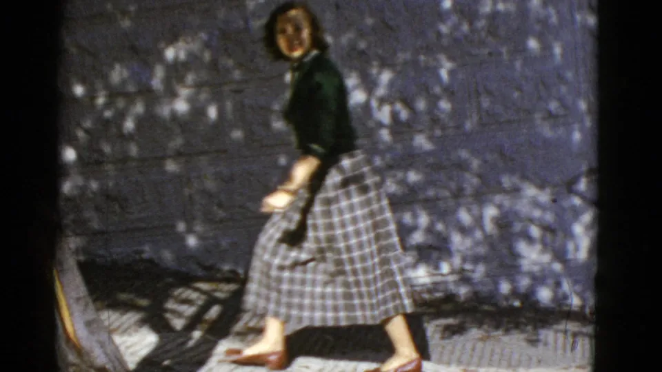 A woman in mid-century clothing walks beside a wall on a sun-dappled San Francisco sidewalk.