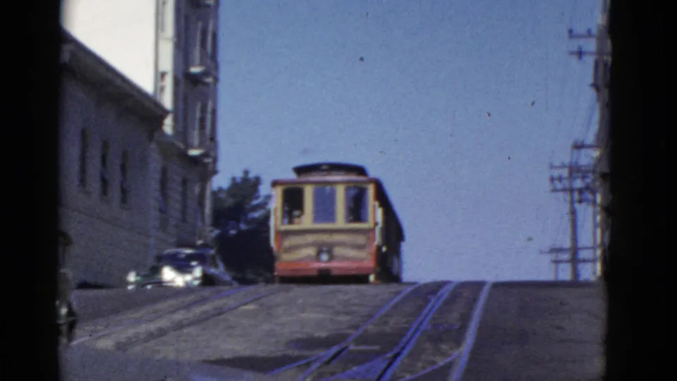 A red-and-cream cable car climbs a steep San Francisco street with tracks, buildings, and utility poles visible.