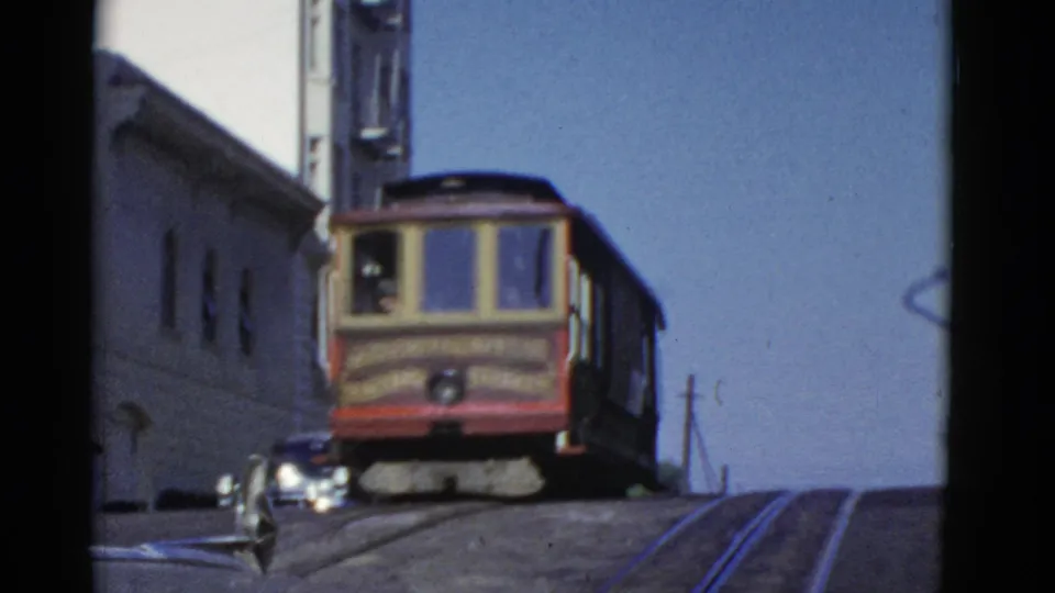 A cable car fills more of the frame as it climbs a steep San Francisco hill past a parked car and street rails.