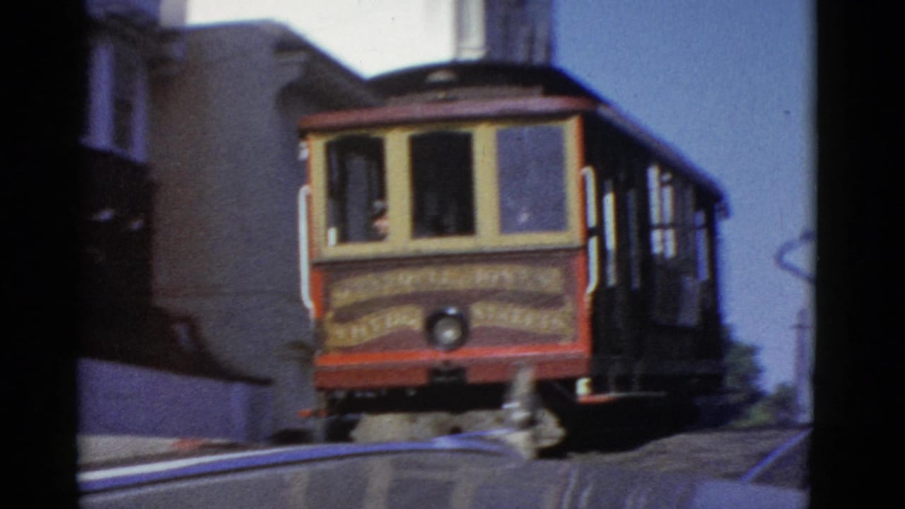 1952 San Francisco home-movie frame showing a red and cream cable car traveling along tracks between city buildings.
