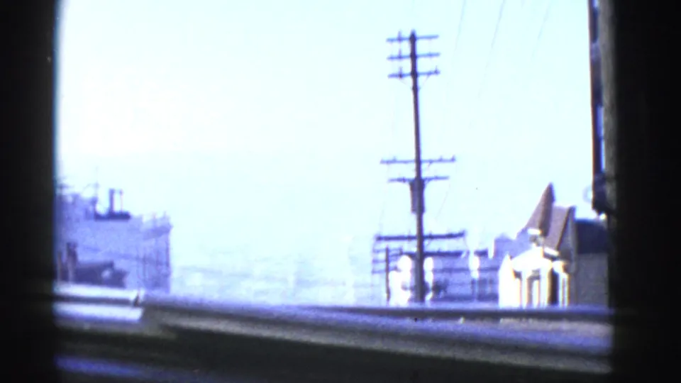 San Francisco home-movie cutaway with utility poles, rooftops, bright haze, and a distant ship or waterfront form.