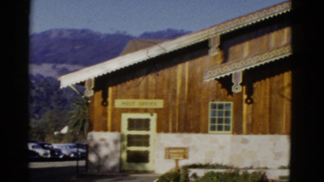 1952 home-movie frame of a rustic wooden post office exterior with decorative trim, stone foundation, parked cars, and hills behind it.