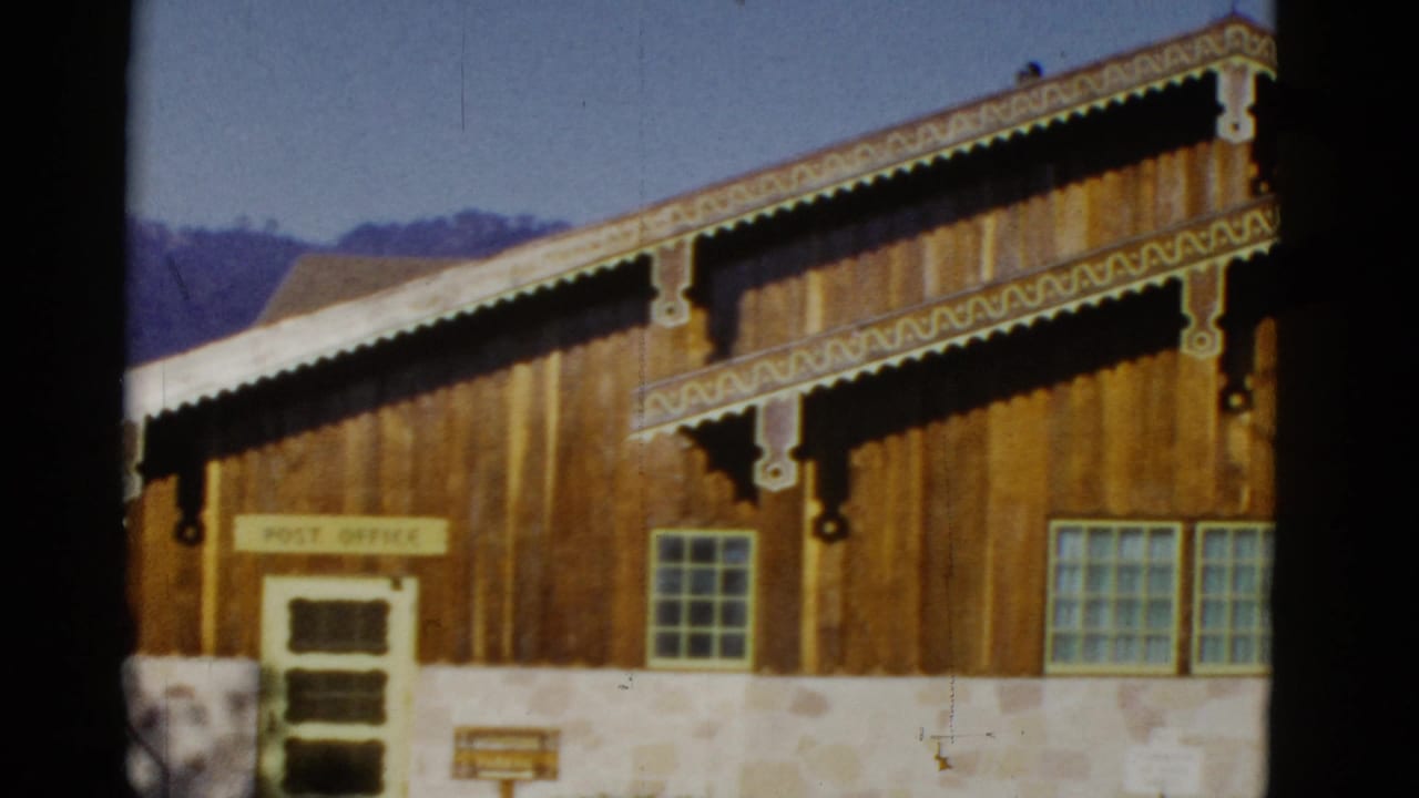 Front of a wood-and-stone post office building with decorative roof trim, green door, windows, and mountain backdrop.