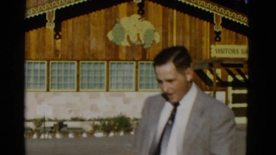 A suited man walks past a rustic wooden building with glass-block windows and a partially visible visitor sign in 1952 footage.