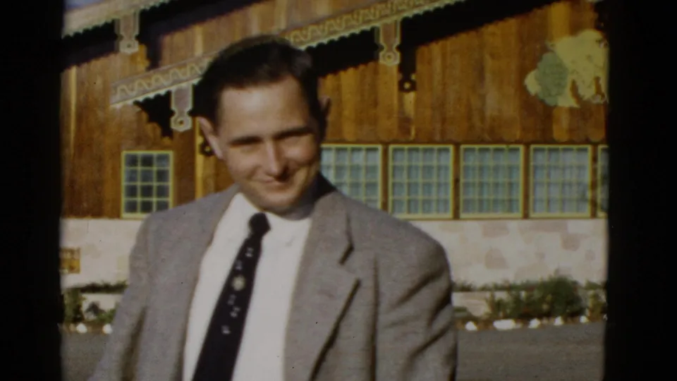A smiling man in a light gray suit faces the camera near the rustic storefront in this 1952 San Francisco home-movie frame.