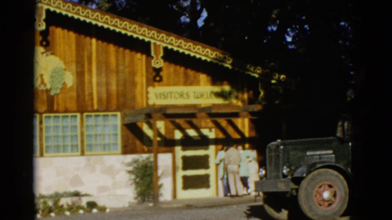1952 8mm frame of a decorated visitors building in California with adults at the doorway and a green truck nearby.