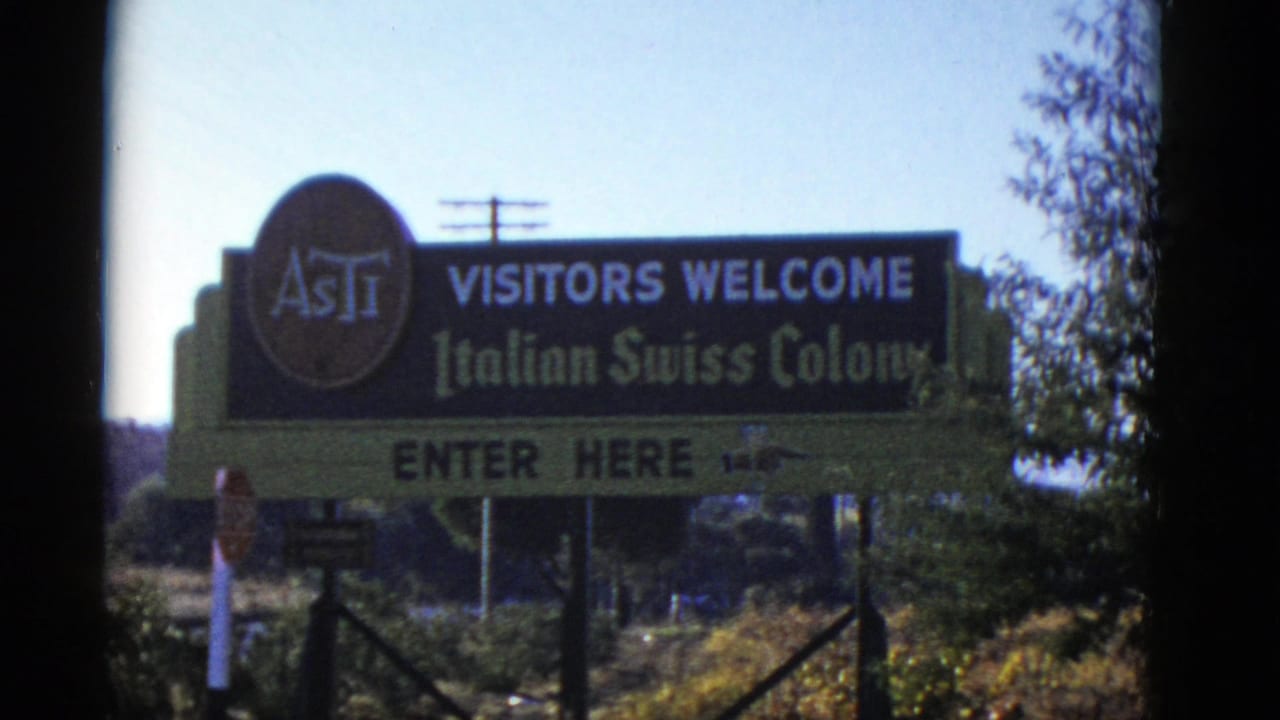 1952 8mm frame showing an Italian Swiss Colony roadside welcome sign with ASTI logo, trees, power lines, and brush nearby.