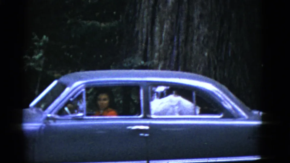 In this 1950s home-movie frame, a woman in a red top is seen through the window of a dark sedan in a wooded California setting.